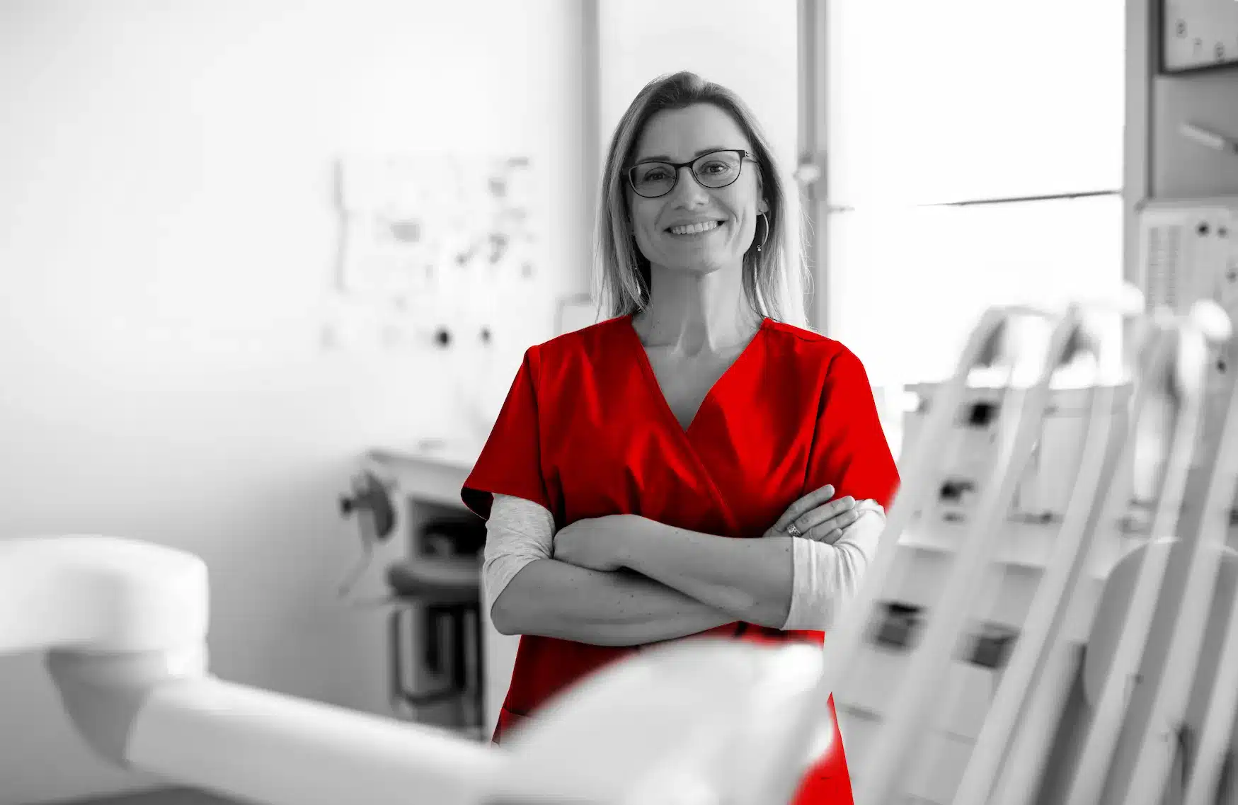 A imagem mostra uma mulher com roupas hospitalares e ela está em um ambiente hospitalar. Ela sorri usa óculos, tem os cabelos médios e olha para a câmera sorrindo, enquanto cruza os braços. A imagem toda está em branco e preto, sendo que somente suas roupas estão em vermelho, em destaque.