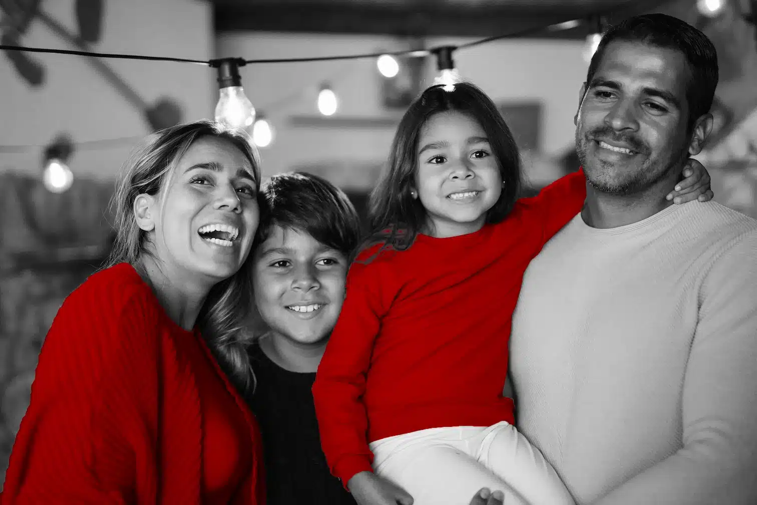A imagem mostra uma família reunida: mãe, pai, um garoto e uma menina. A mãe está do lado do filho, sorrindo, enquanto o pai segura a filha no colo. Os quatro possuem expressões alegres. A imagem está em branco e preto, sendo que somente as blusas da mãe e da filha aparecem em vermelho, em destaque.
