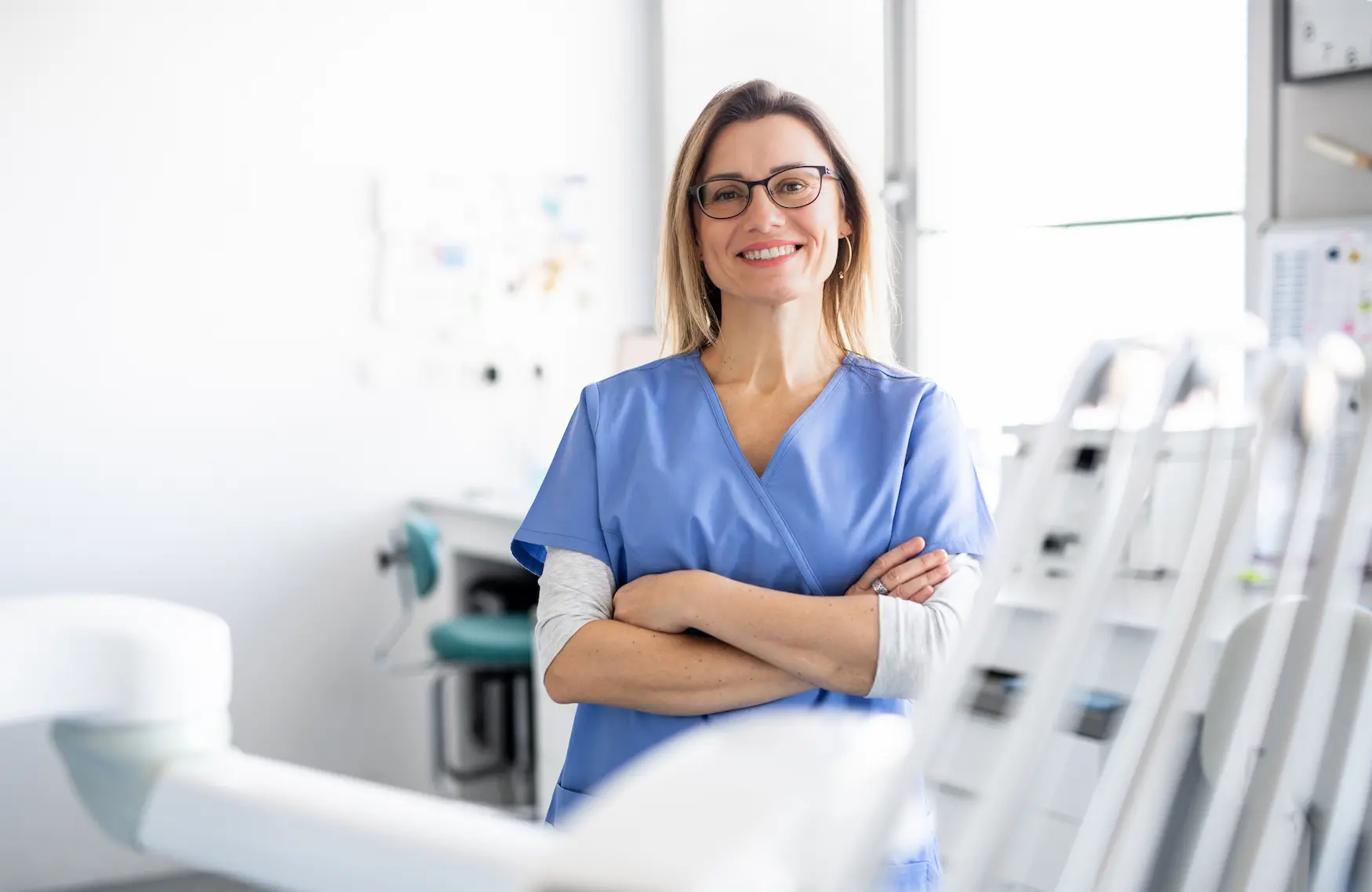 A imagem mostra uma mulher com roupas hospitalares e ela está em um ambiente hospitalar. Ela sorri usa óculos, tem os cabelos médios e olha para a câmera sorrindo, enquanto cruza os braços.