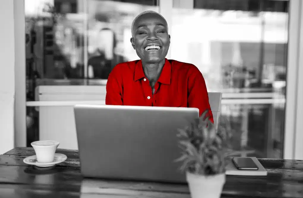 A imagem ostra uma mulher negra, sorrindo para a câmera. Ela está sentada em uma cadeira, perto de uma mesa. Na mesa, há um laptop aberto, uma xícara de café, um caderno, um smartphone e um vaso com plantas. Toda a imagem está em branco e preto, sendo que somente a blusa da mulher aparece em vermelho, em destaque.