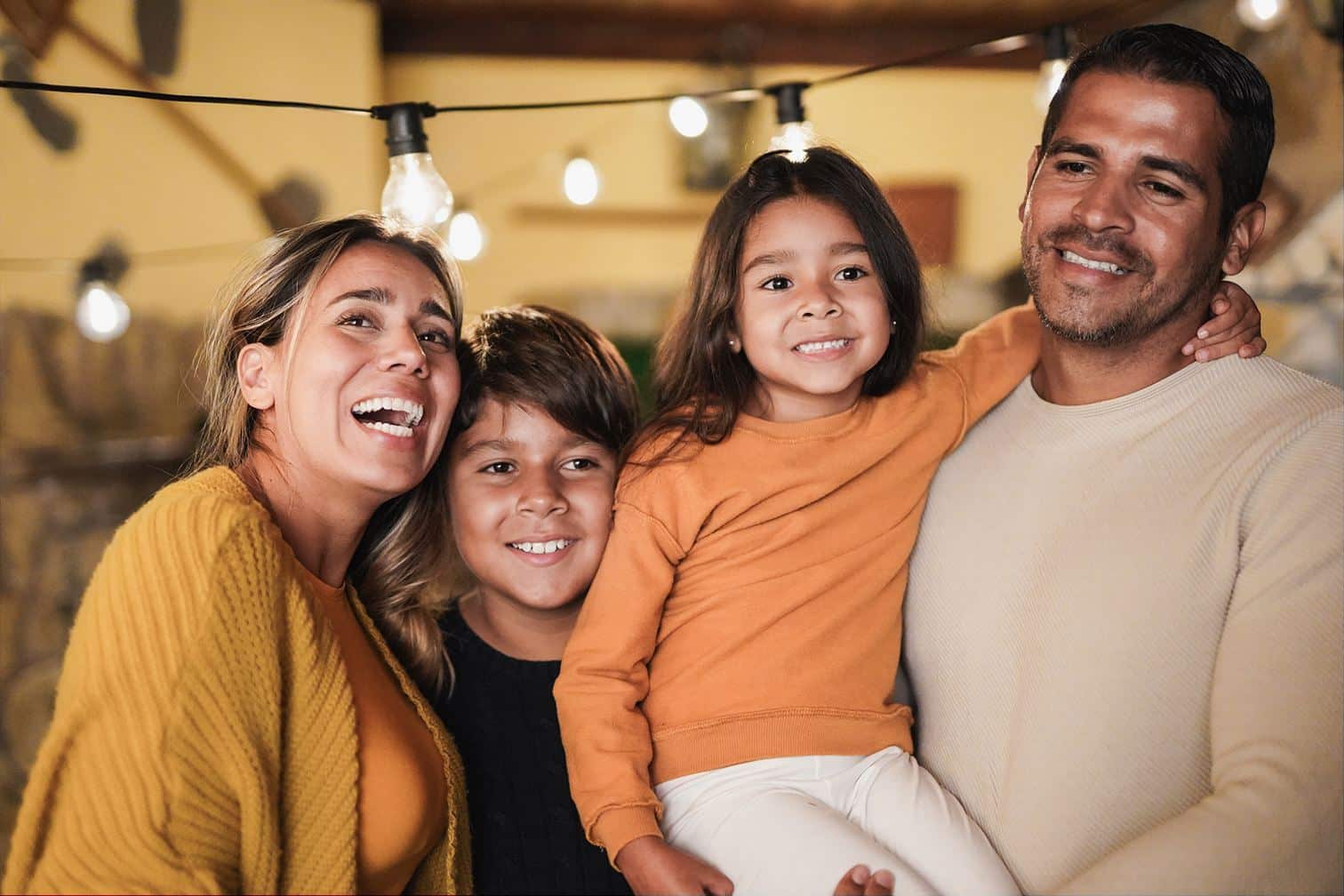 A imagem mostra uma família reunida: mãe, pai, um garoto e uma menina. A mãe está do lado do filho, sorrindo, enquanto o pai segura a filha no colo. Os quatro possuem expressões alegres.