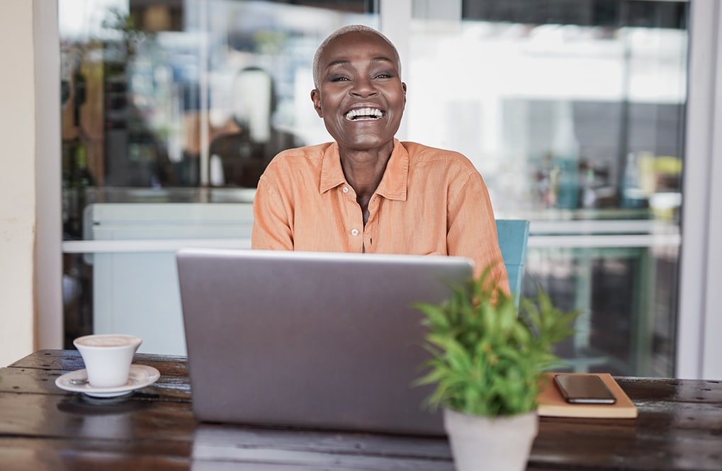 A imagem ostra uma mulher negra, sorrindo para a câmera. Ela está sentada em uma cadeira, perto de uma mesa. Na mesa, há um laptop aberto, uma xícara de café, um caderno, um smartphone e um vaso com plantas.